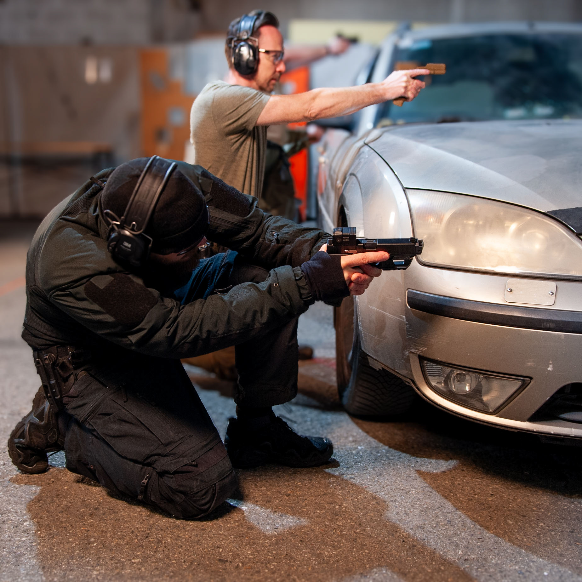 Zwei Sportschützen sind hinter einem Trainingsfahrzeug in Deckung gegangen, Der vordere kniet.